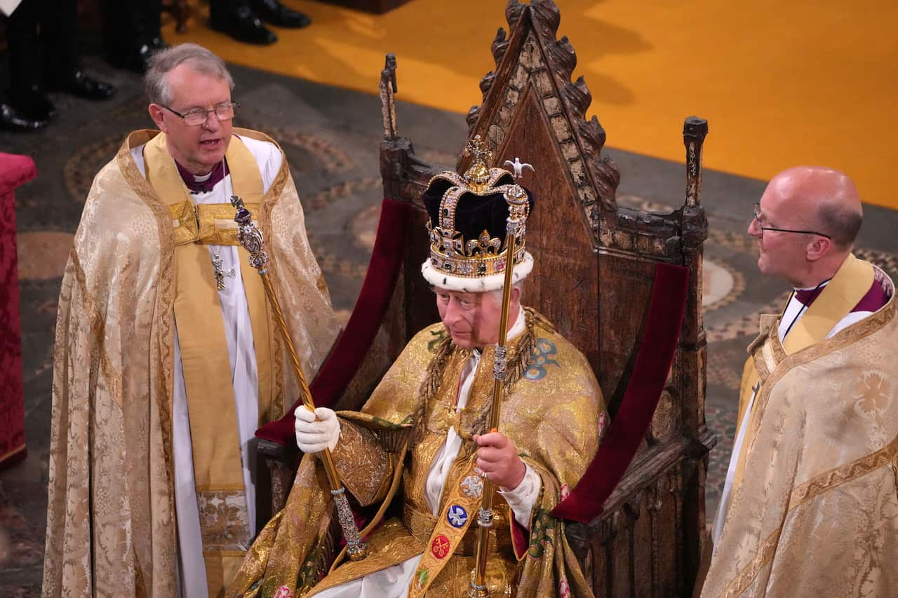 King Charles, wearing a robe with gold sleeves, sits on a throne after his coronation. He holds sceptres in both hands, flanked by two men in ceremonial robes on either side.