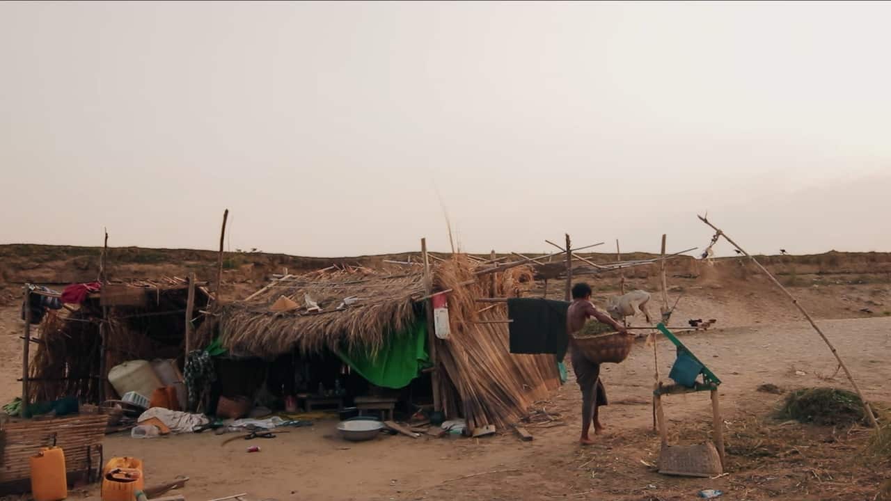 A makeshift shelter stands on a barren, sandy landscape. Nearby, a person carries a basket beside a simple wooden structure, while a goat and several chickens roam in the background