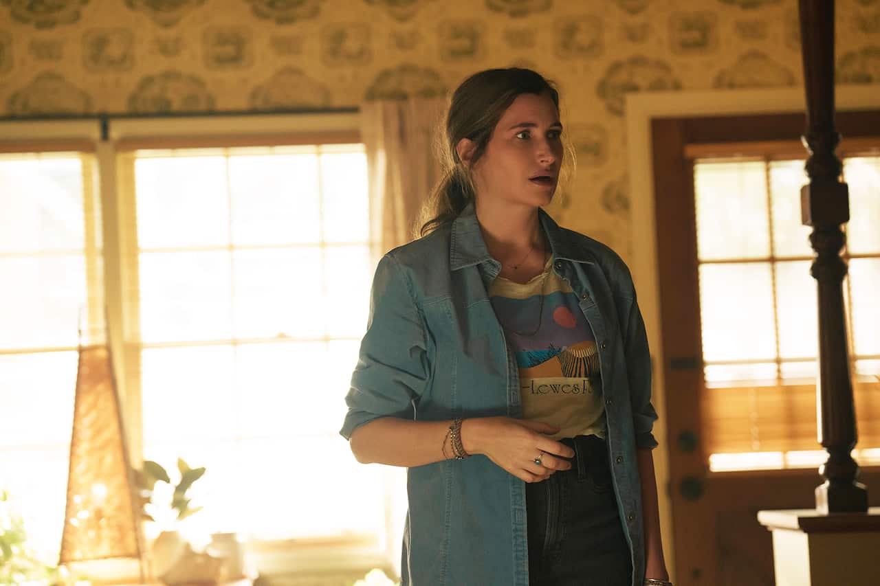 A woman in a denim shirt over a T-shirt stands in front of a sun-lit window with a worried expression.