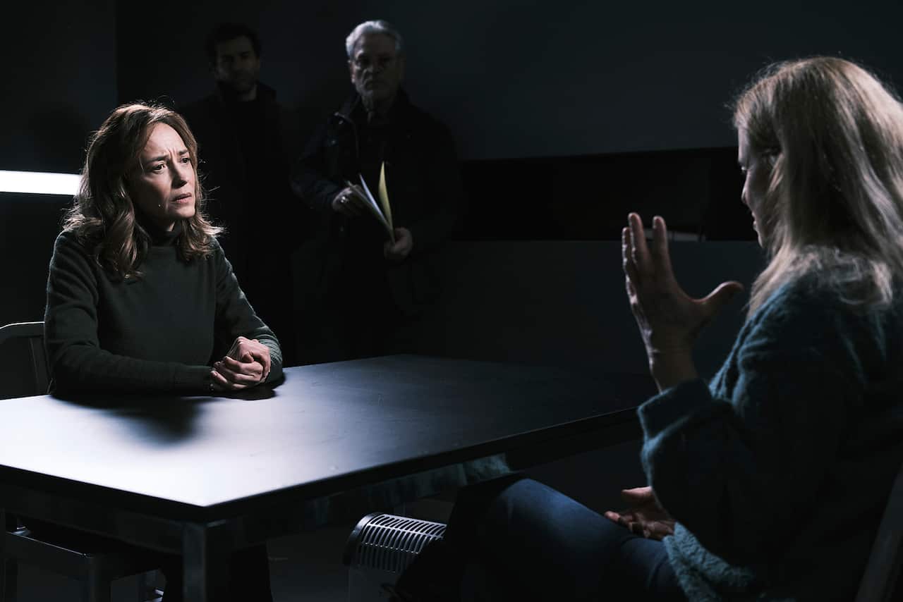 A woman sits at a table, looking earnestly at another woman sitting opposite her, who is only seen from side-on. A white-haired man holding a folder can just be discerned in the background. 