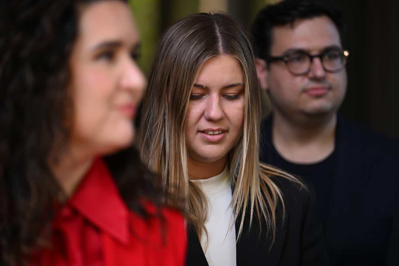 A woman standing between another woman and a man with her head tilted slightly down.