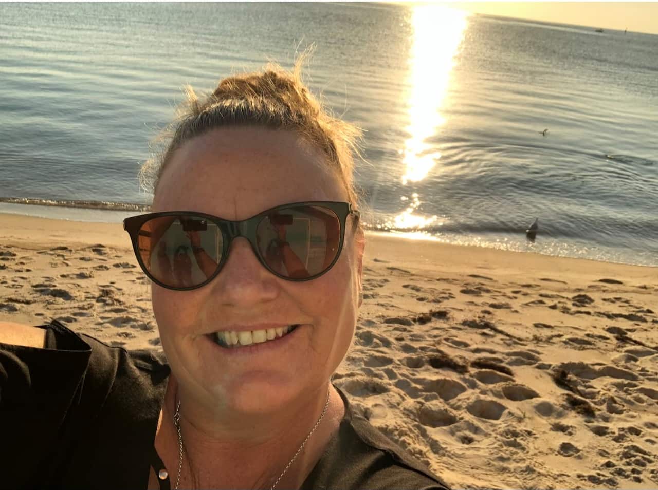 A woman smiles with sunglasses in front of a beach.