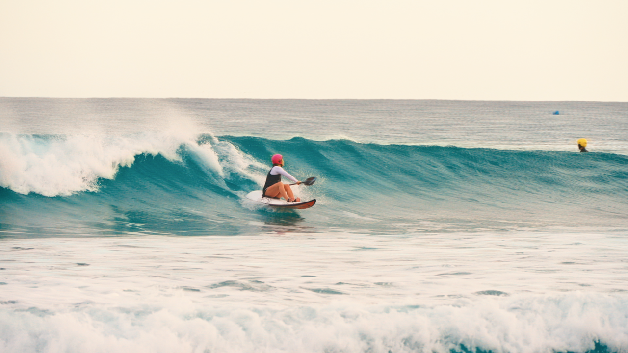 A woman on a waveski in a bright helmut rides a wave, paddle in hand. 