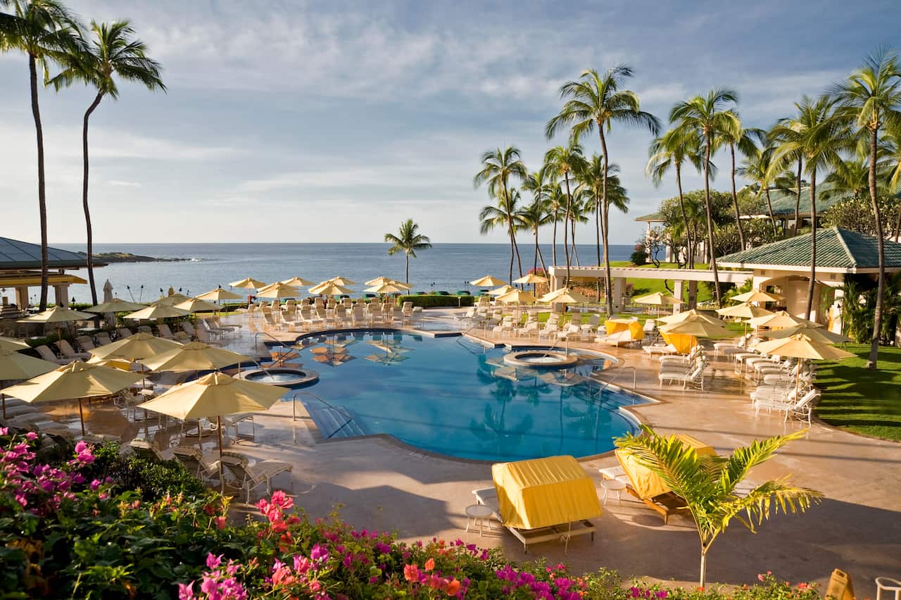 A resort pool in front of the ocean, surrounded by palm trees and deck chairs 