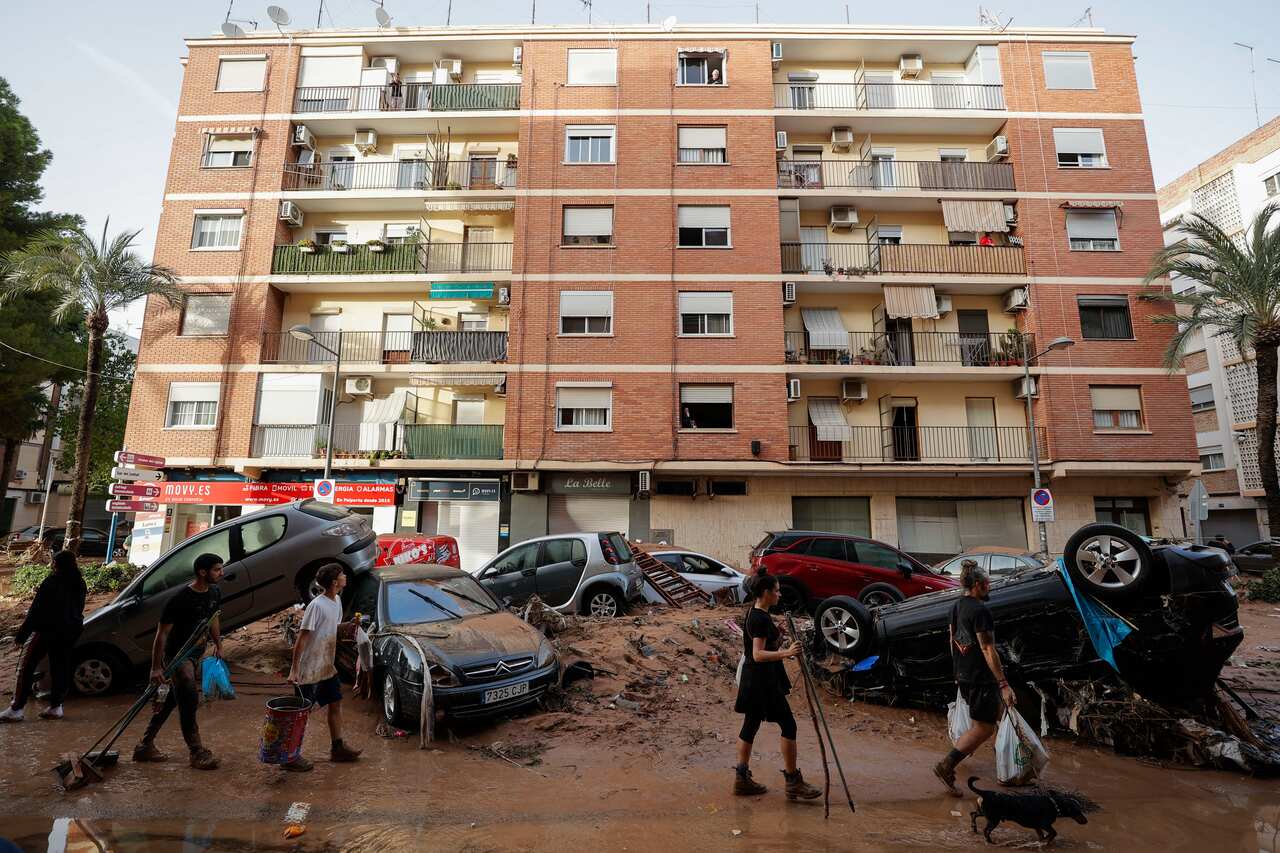 A group of people walk past cars and muddy debris in the street in front of an apartment block 