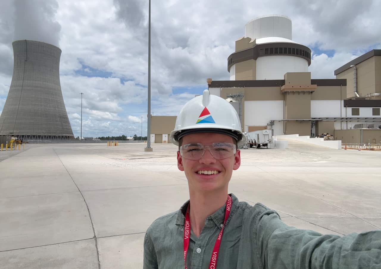 A young man in a hard hat and safety glasses smiles in front of a nuclear power plant