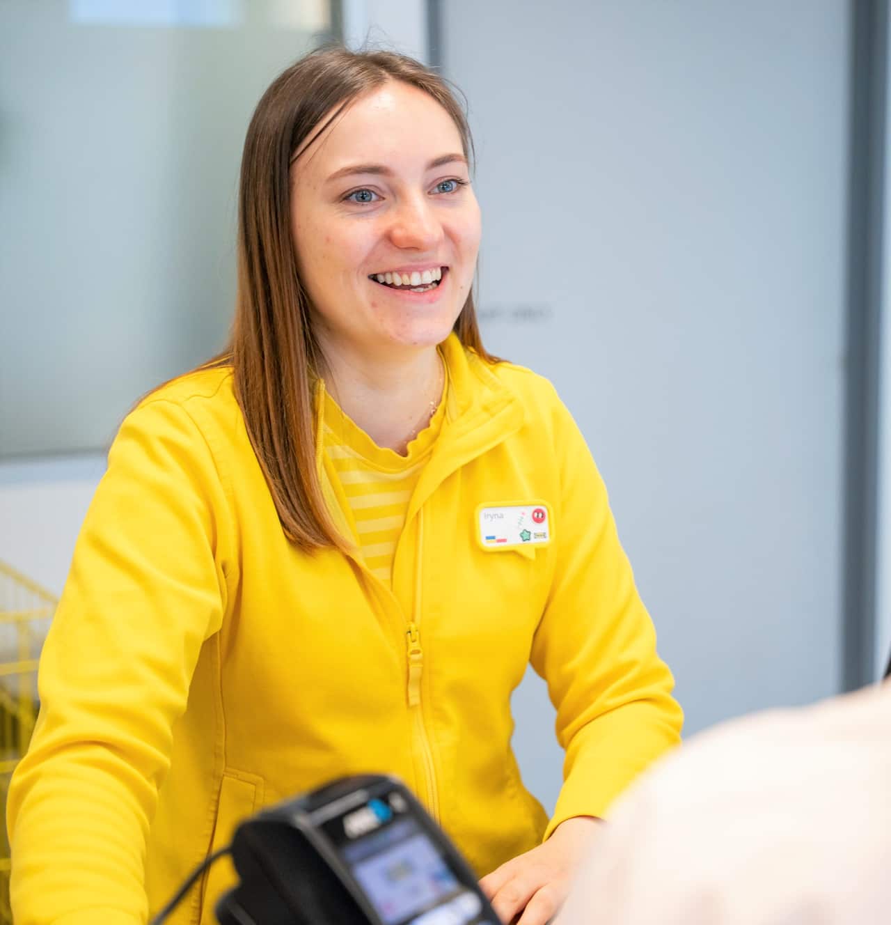 A young woman with brown hair and wearing a yellow jacket stands at a service counter.