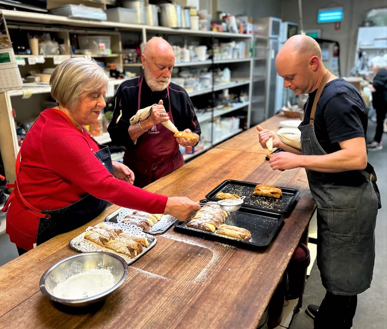 A woman in a red jumper stands at a bench with two men, filling Italian pastries.