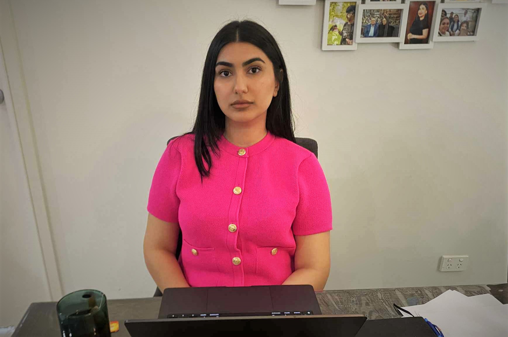 A young woman of Iranian descent, with long straight dark hair, wearing a bright pink short-sleeved top with gold buttons, sitting at a desk in front of a laptop. Some family photos hang on the wall behind her.