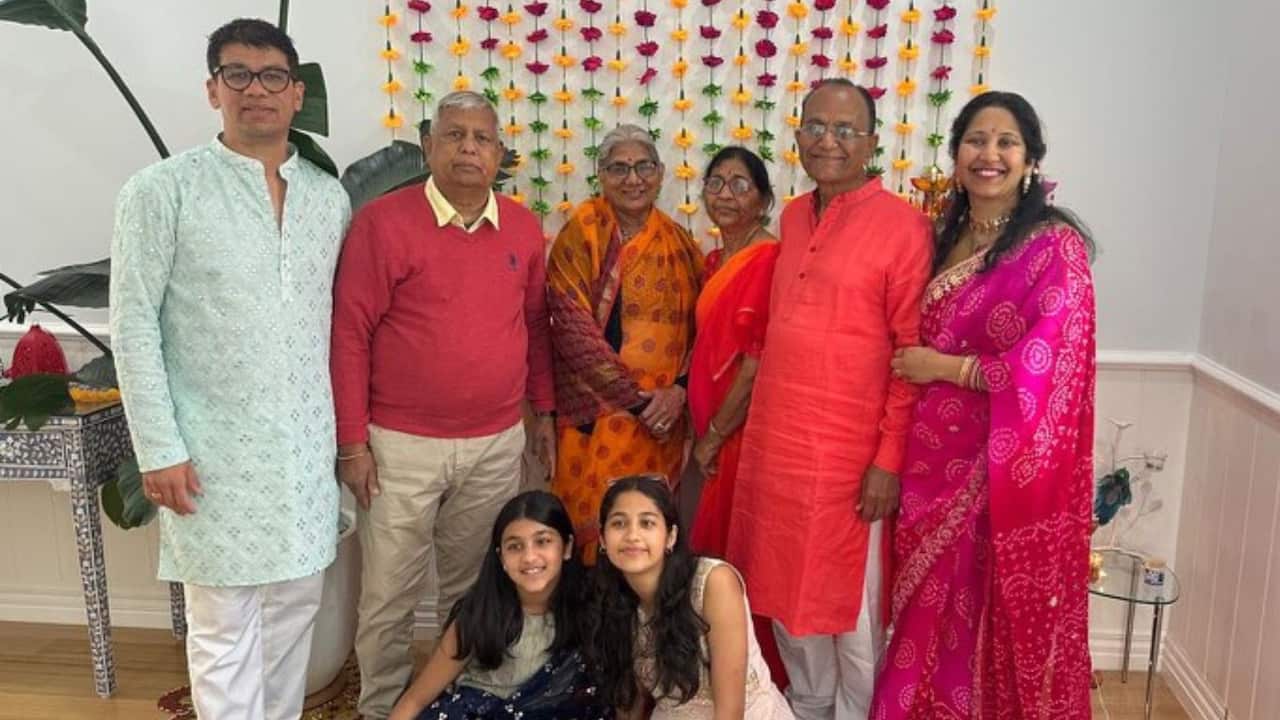 A group of eight people pose inside for a picture. All are wearing traditional Indian dress. A man and a woman are standing either side of two older men and two older women. Two girls are sitting in front of them