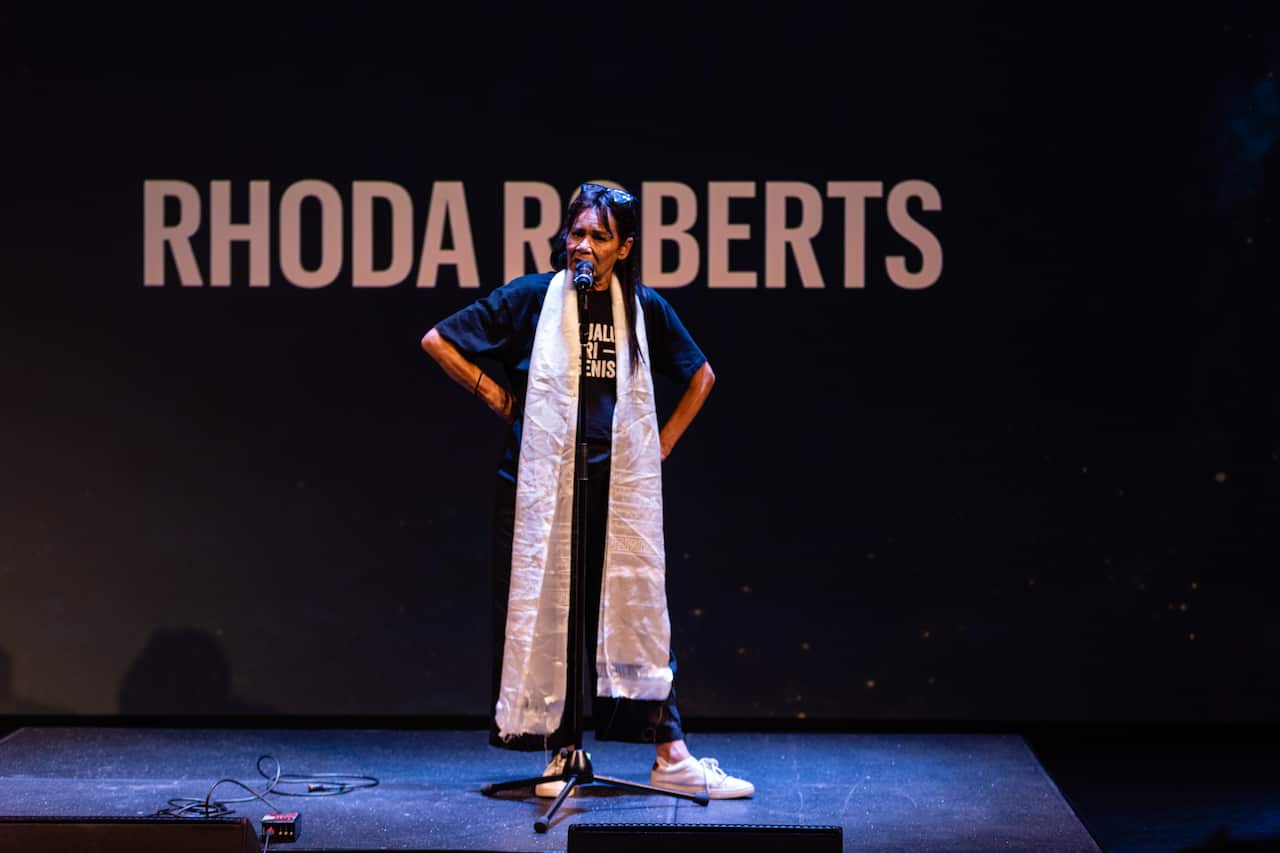 A woman speaking while standing on a stage. She is standing in front of a screen that says: RHODA ROBERTS.