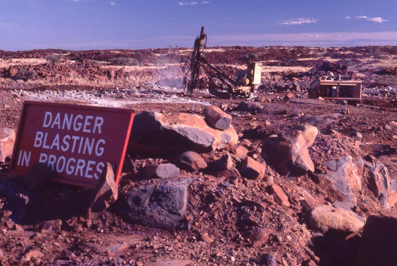 A red sign reading Danger blasting in progress sitting on a rocky mining landscape with drilling equipment operating in the background.