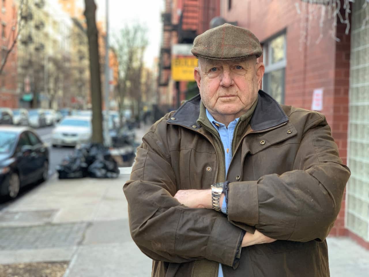 An older man wearing a brown jacket and flat cap standing on the street with his arms crossed and looking into the camera