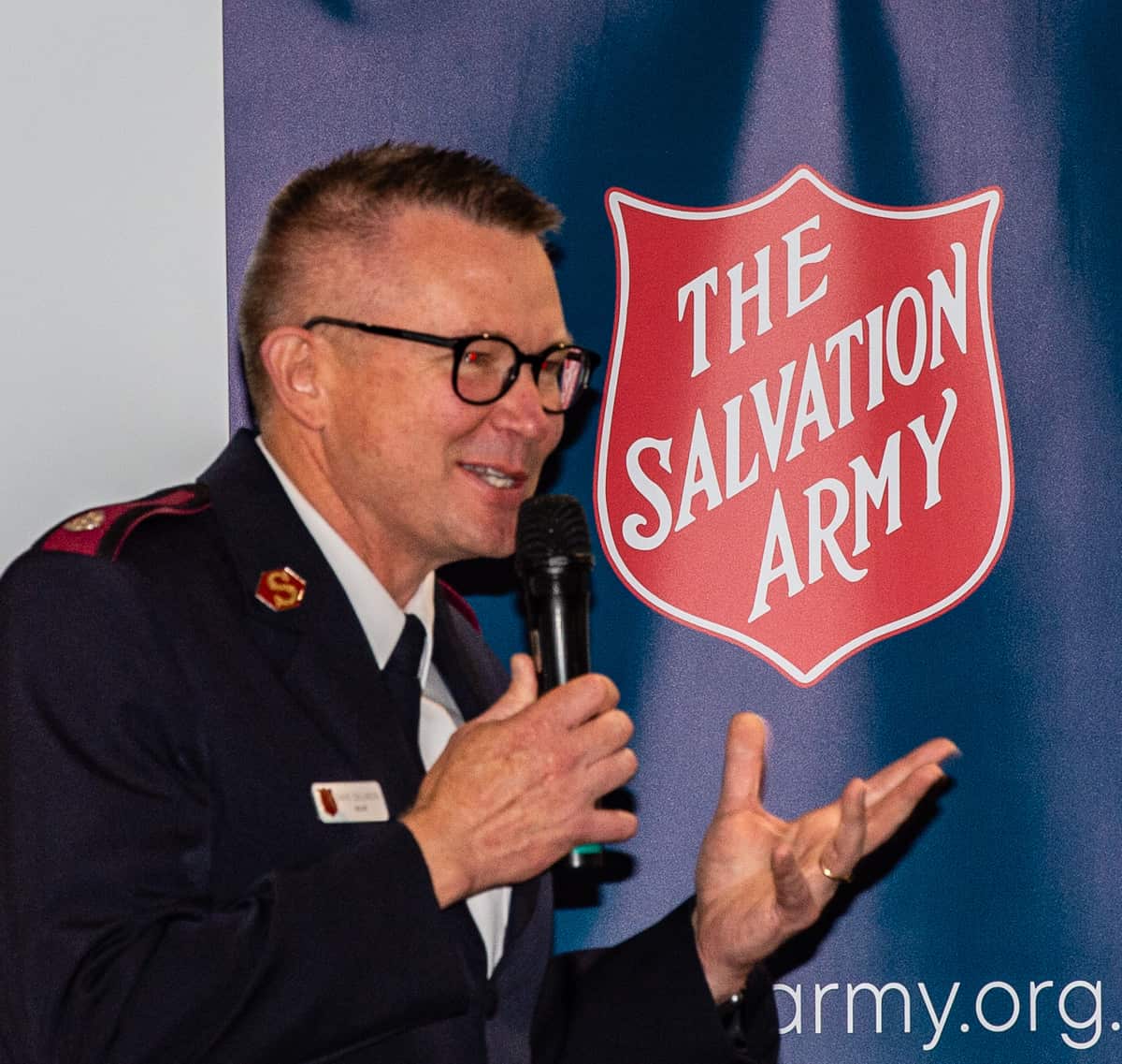 Major David Collinson speaking into a microphone in front of a Salvation Army shield.