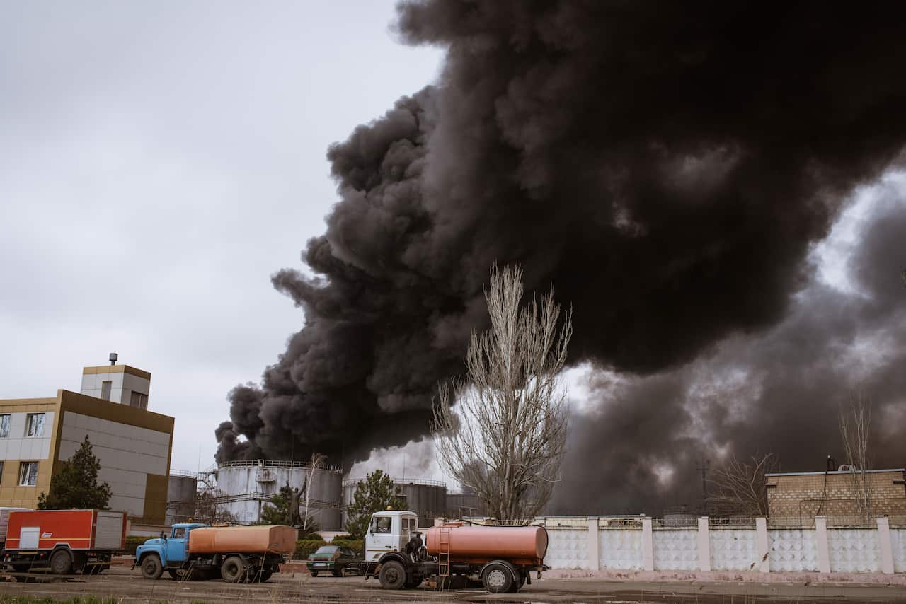 Three trucks are seen parked on a road as black smoke is seen in the background.