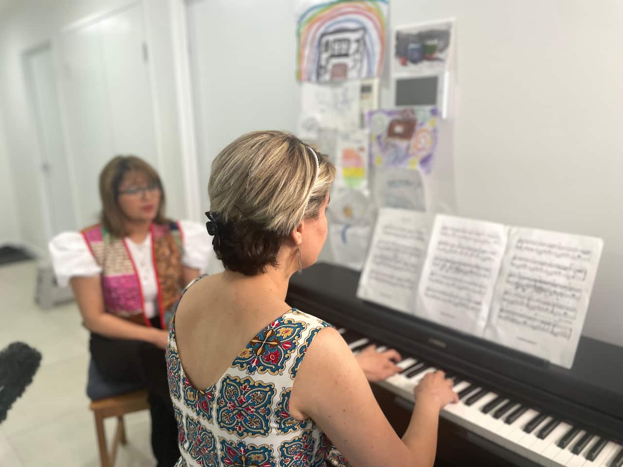 A woman plays a piano with another woman on her left watching.