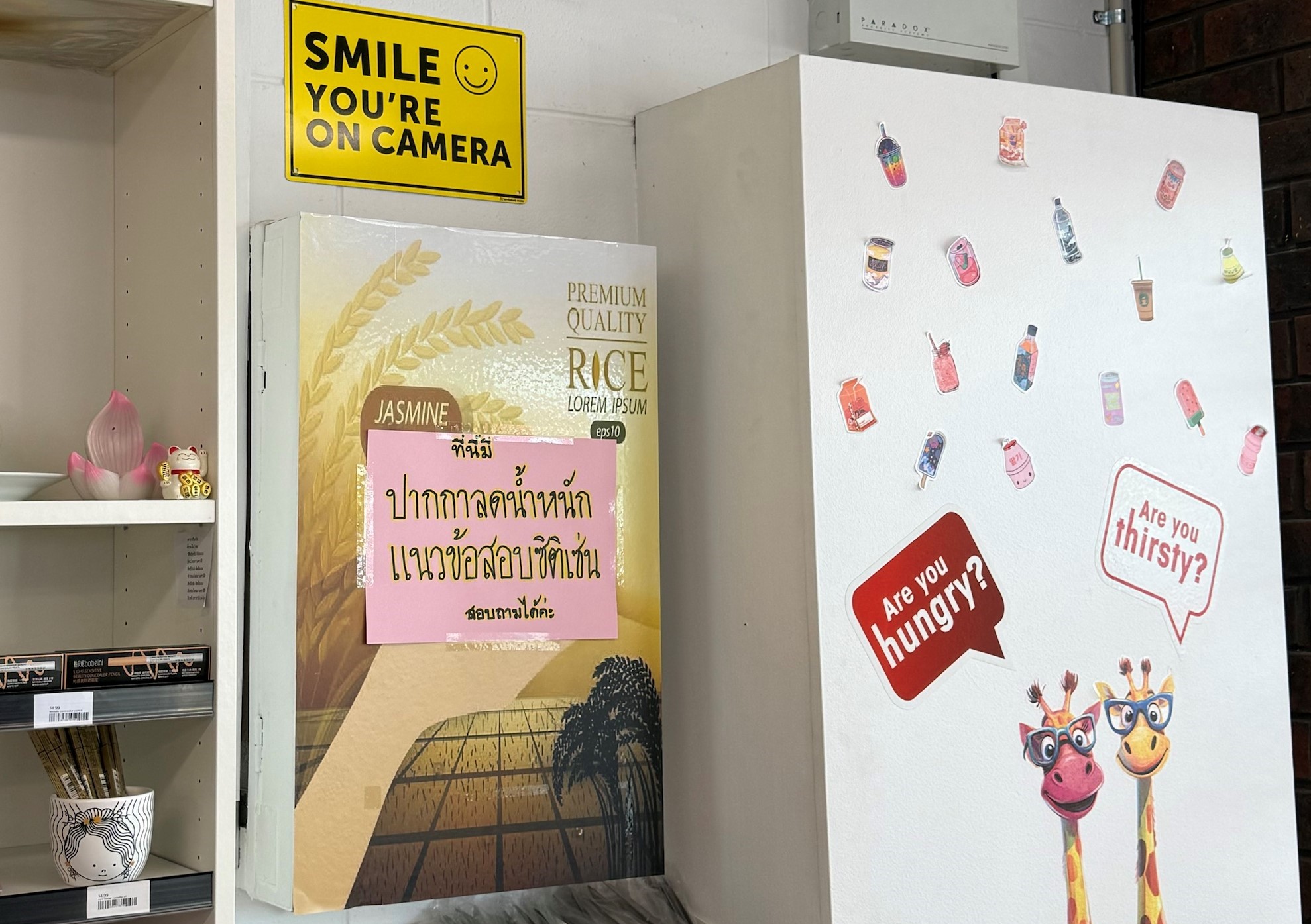 A Thai poster is stuck on another poster on a grocery store wall. A white panel is attached to the wall next to the poster, plastered with colourful stickers.