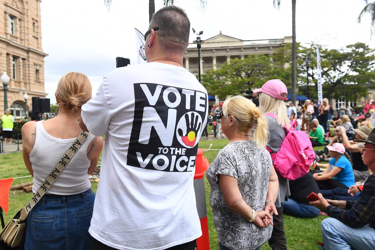 Four people standing in front of a building with their backs turned. One has a shirt that says Vote no to the Voice