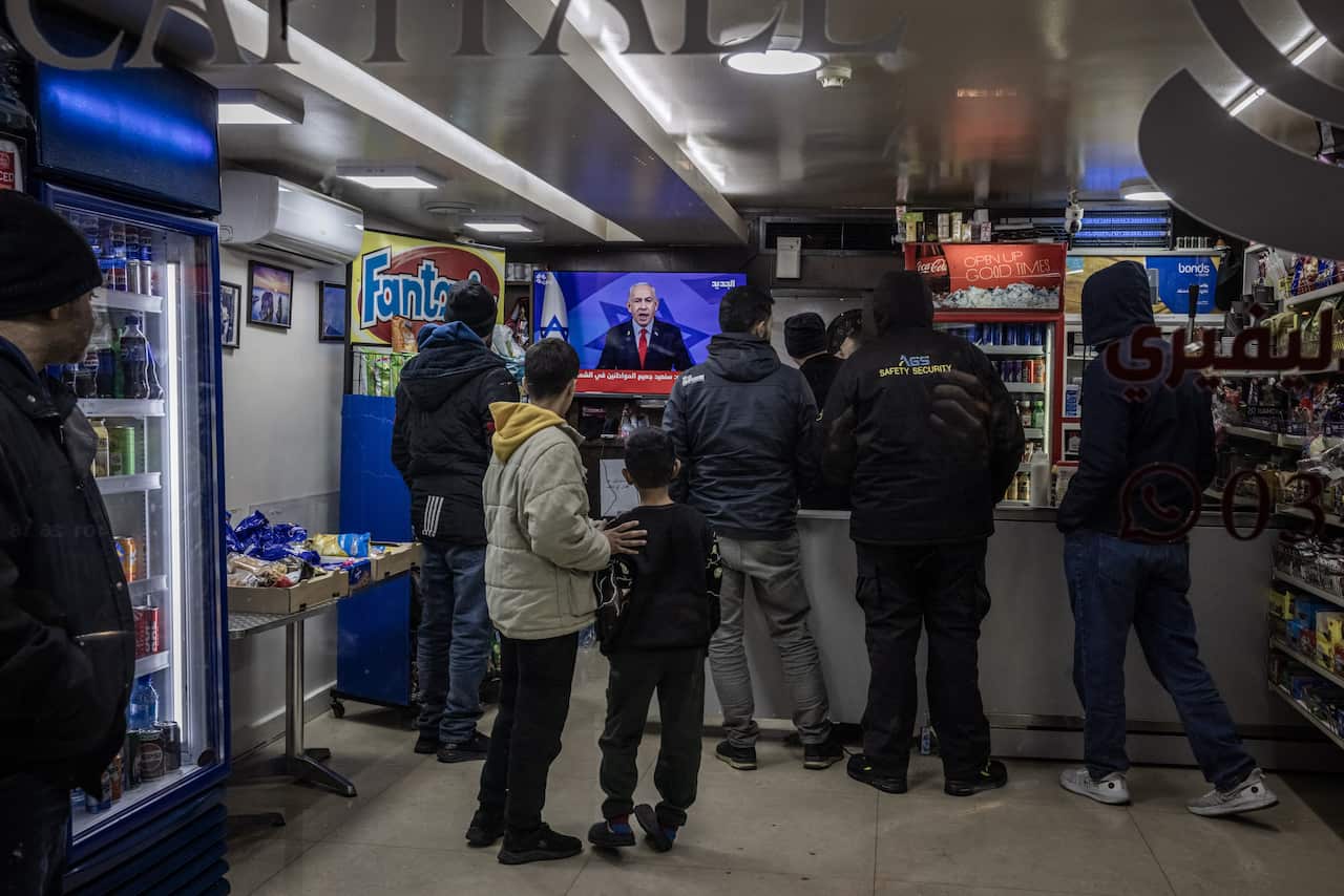 People are standing inside a shop, watching a television playing the news.
