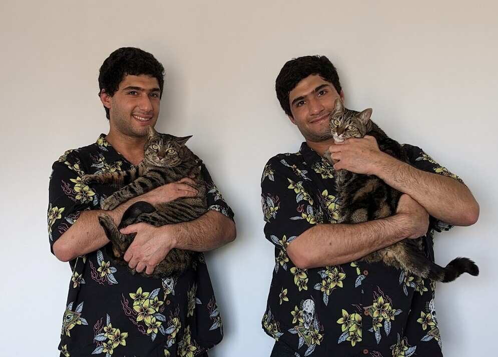 Two identical brothers wearing the same shirts, smile as they hold up near identical cats.