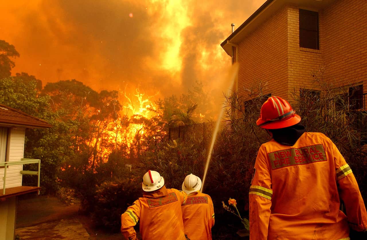 A group of three firefighters wearing orange outfits standing in front of a large blaze. One person is spraying water from a fire hose onto the side of a two-storey brick house.
