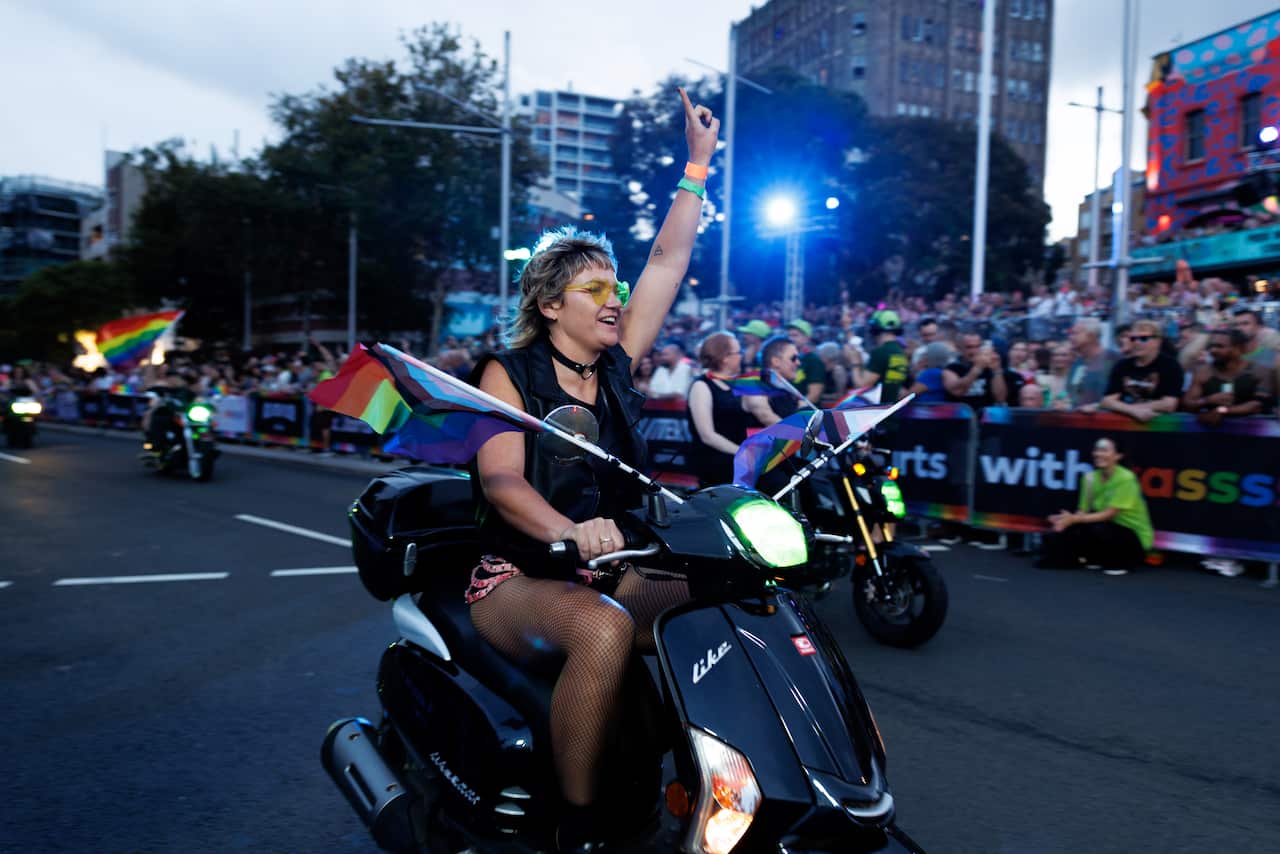 A person with blonde hair and yellow sunglasses rides a black scooter down a city street at dusk. They have one arm raised in a "number one" gesture, and rainbow flags are attached to the front of the scooter.