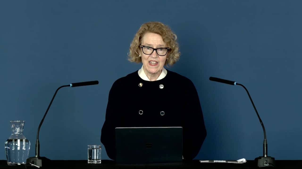 A woman with blonde curly hair and glasses wearing a black top speaks behind a lectern flanked by two podium microphones.