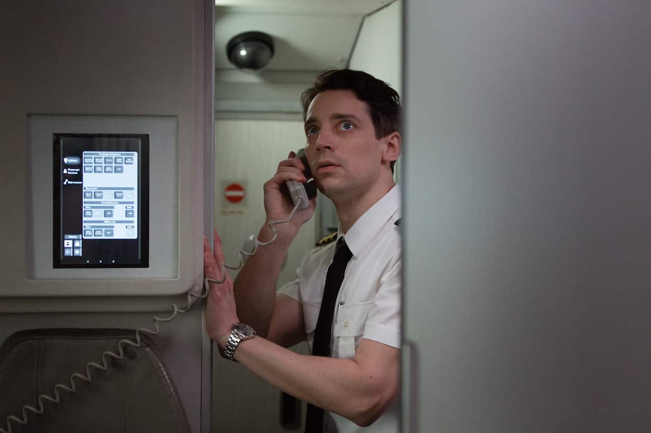 A worried looking man in a pilot's uniform talks on a phone on a plane.