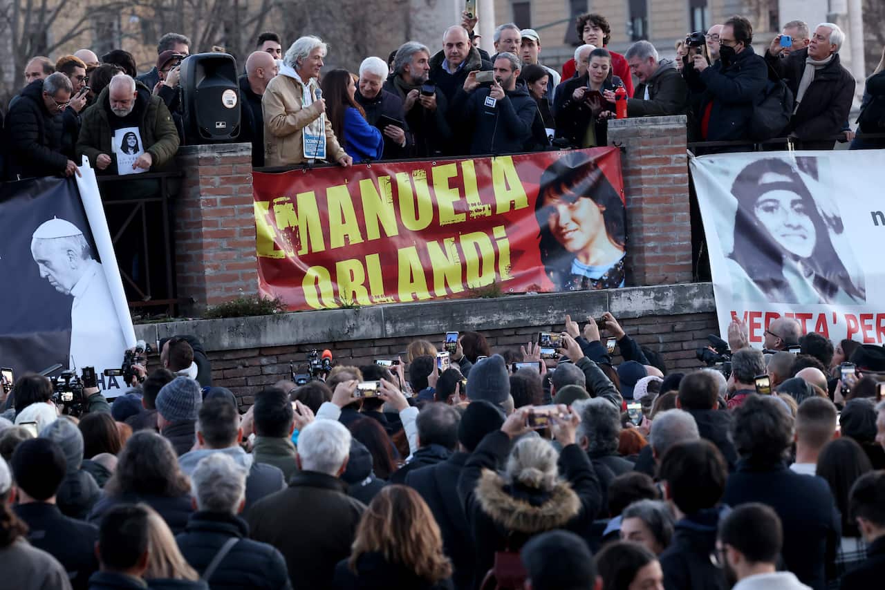 A crowd of people in winter down jackets look up at a speaker with a microphone who stands in front of a sign that reads, 'Emanuela Orlandi'.