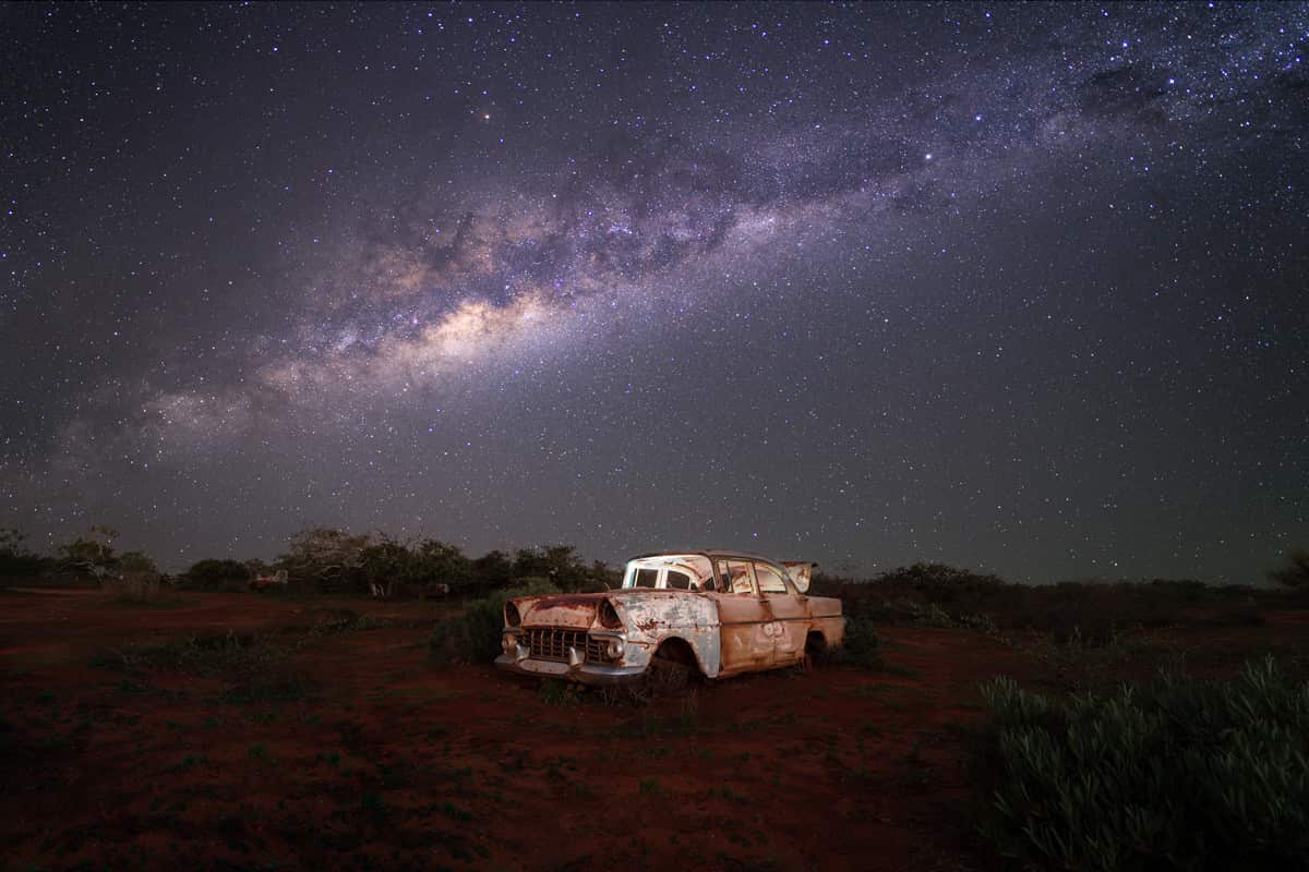 An abandoned old car in the bush with the stars in the night sky visible above.