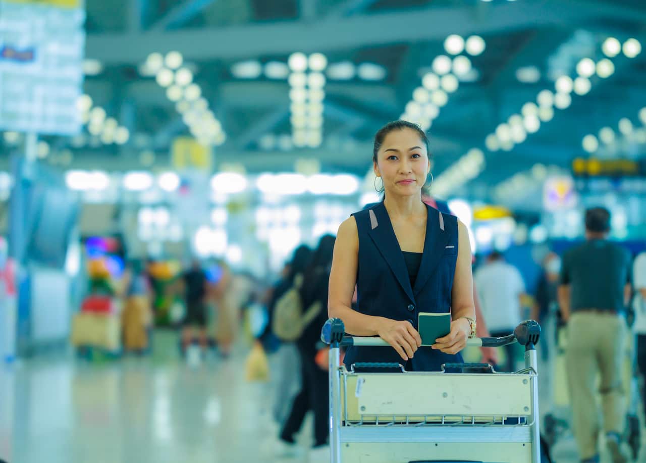 Happy Asian woman holding passport in her hand and standing with her arms on a luggage trolley in the airport.
