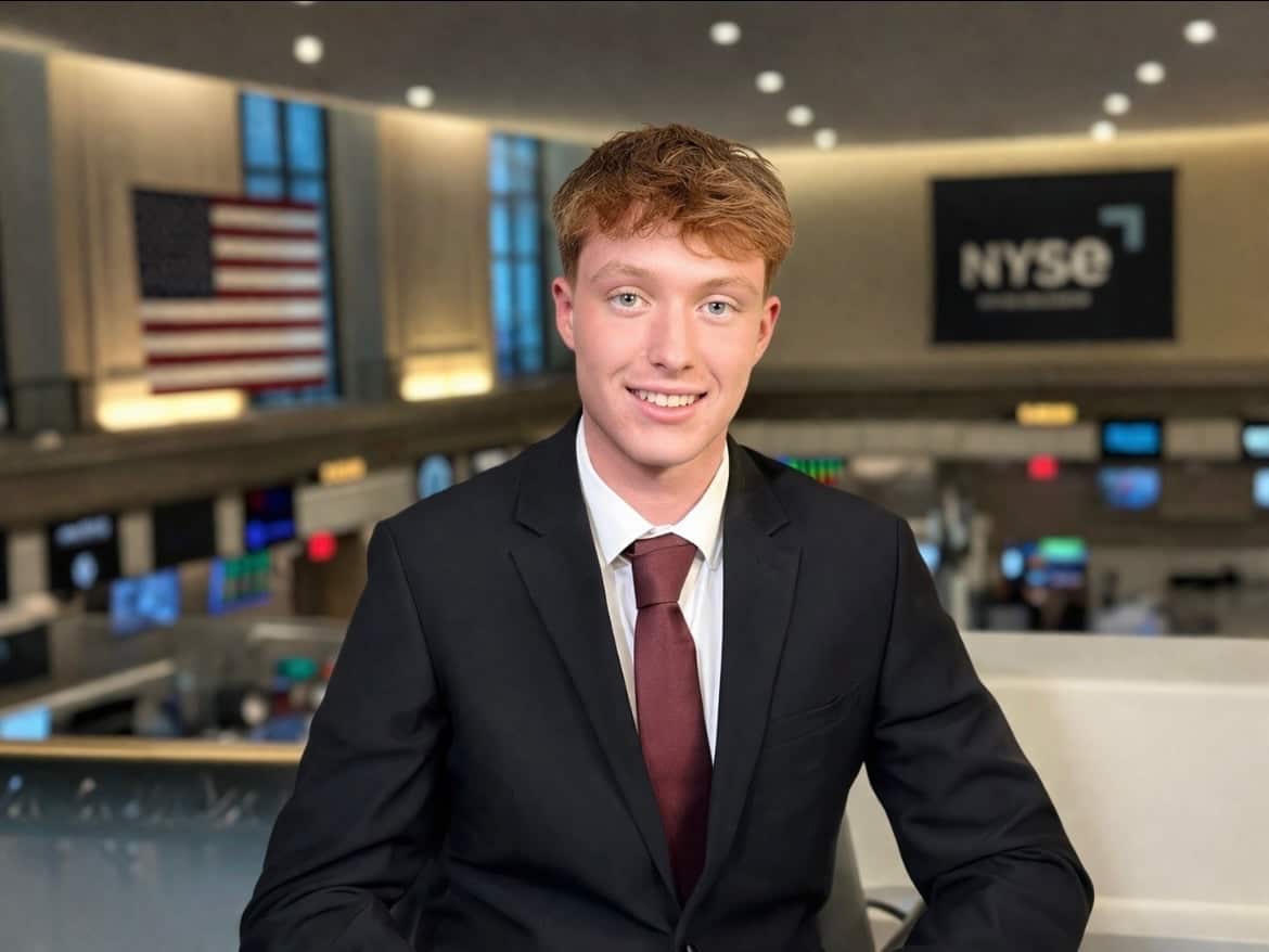 A young man in a suit and tie. In the background is a stock exchange trading floor and the words NYSE on a banner on a wall. A large US flag is hanging in front of another wall.