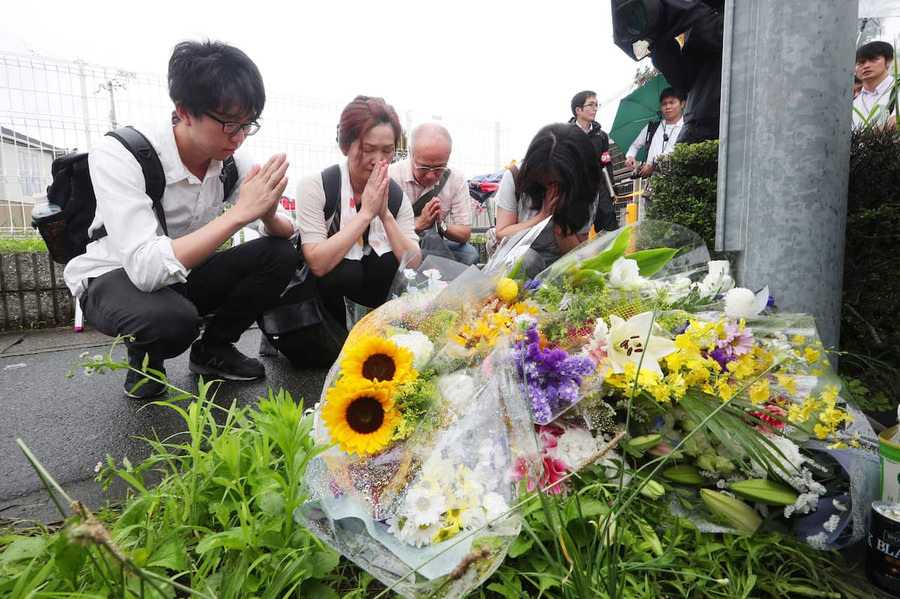 A group of people wearing white shirts clasp their hands in prayer in front of bouquets of flowers. 
