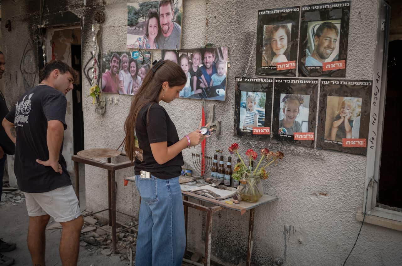 Two people stand in front of a memorial for October 7 victims, which has pictures, flowers and other items. The woman is lighting a candle.