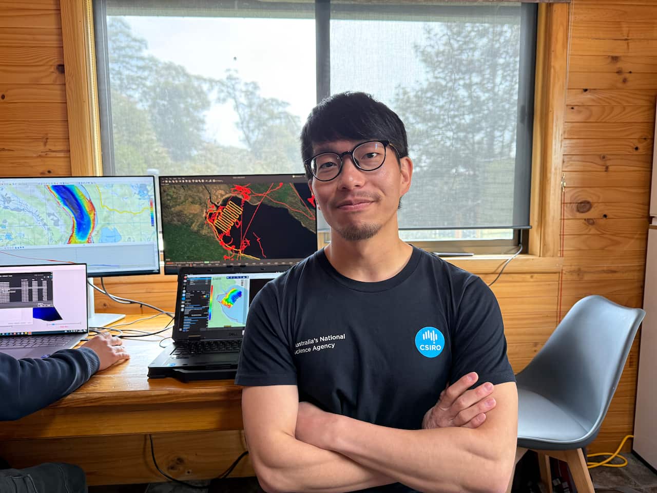 An Asian man with glasses with his arms crossed and smiling. He's seated in front of computers showing maps and diagrams of a lake.