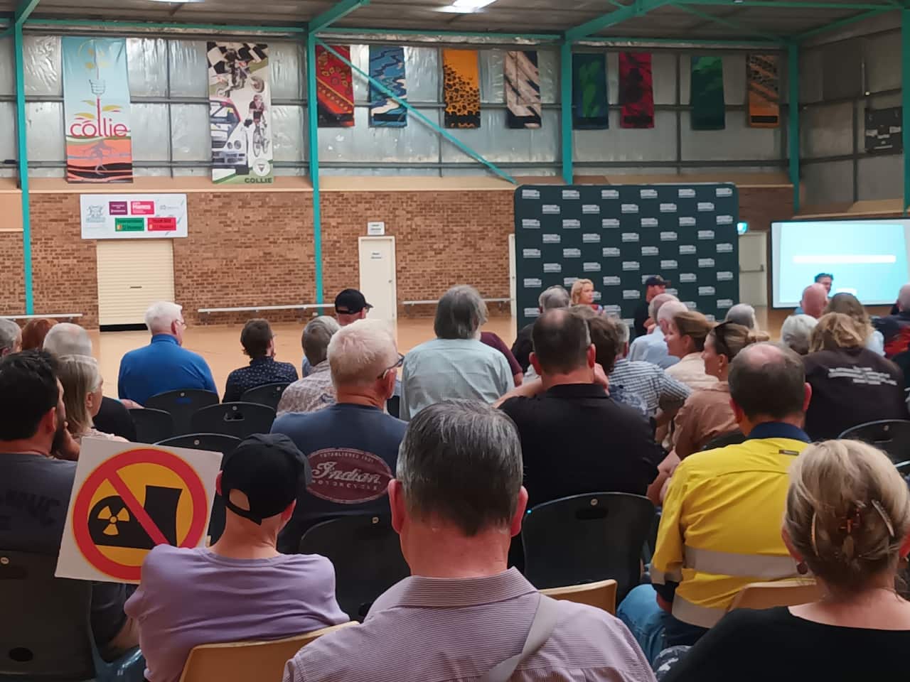 An audience sits in rows of folding chairs facing three speakers at the front of the room in front of a green backdrop