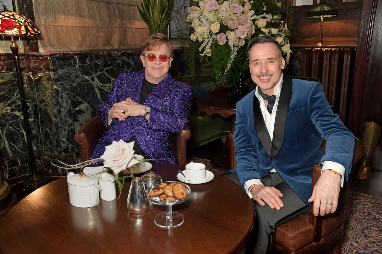 A man in a purple jacket and glasses sits beside a man in a blue jacket, with tea set out on a table in front of them.