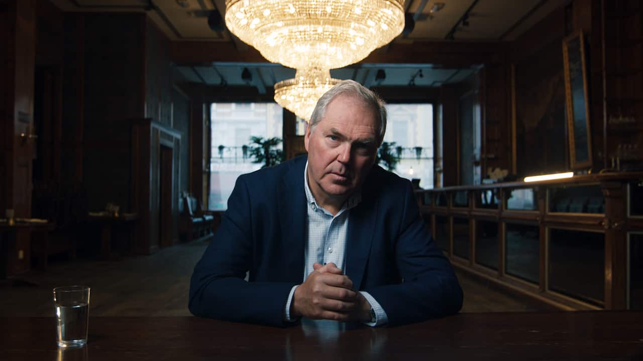 A man in a blue jacket sits at a table, in a dark room. There is a window behind him, and a gold light fitting hanging above him. He leans on the table, hands clapsed in front of him, with a serious expression on his face. 