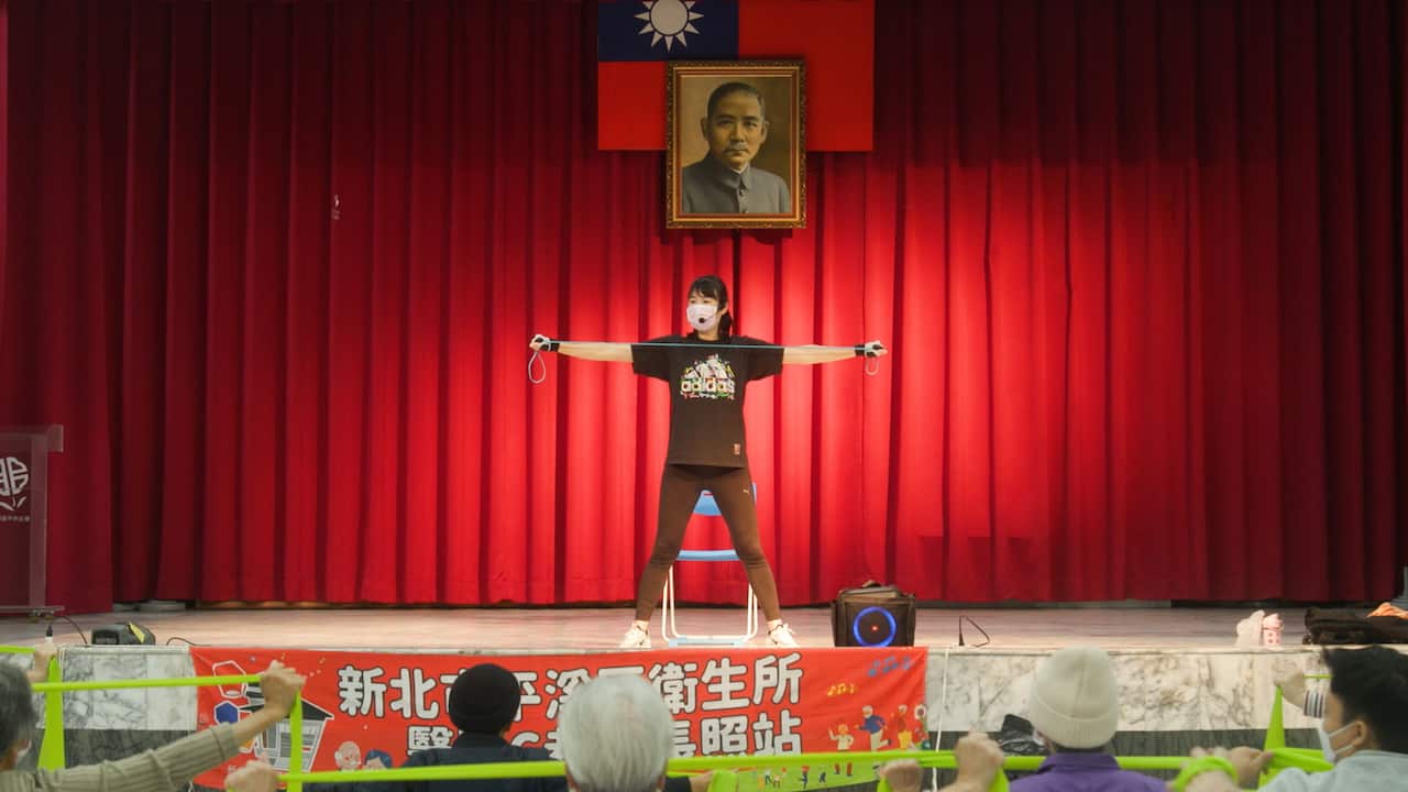 A young woman wearing a medical mask and a microphone is demonstrating an exercise with a resistance band on stage. A few people in the audience are visible, stretching resistance bands with their arms 