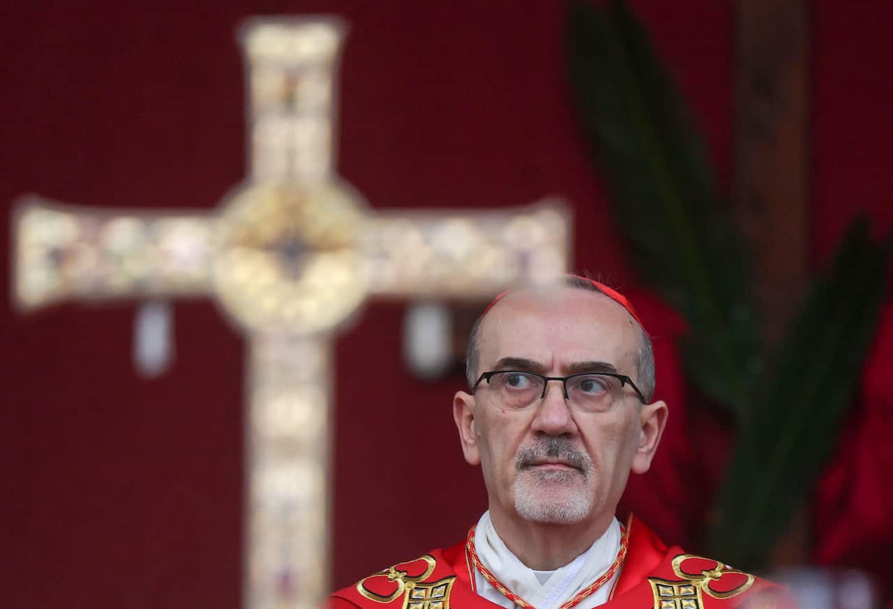 A priest wearing glasses looks on with a cross seen in the background