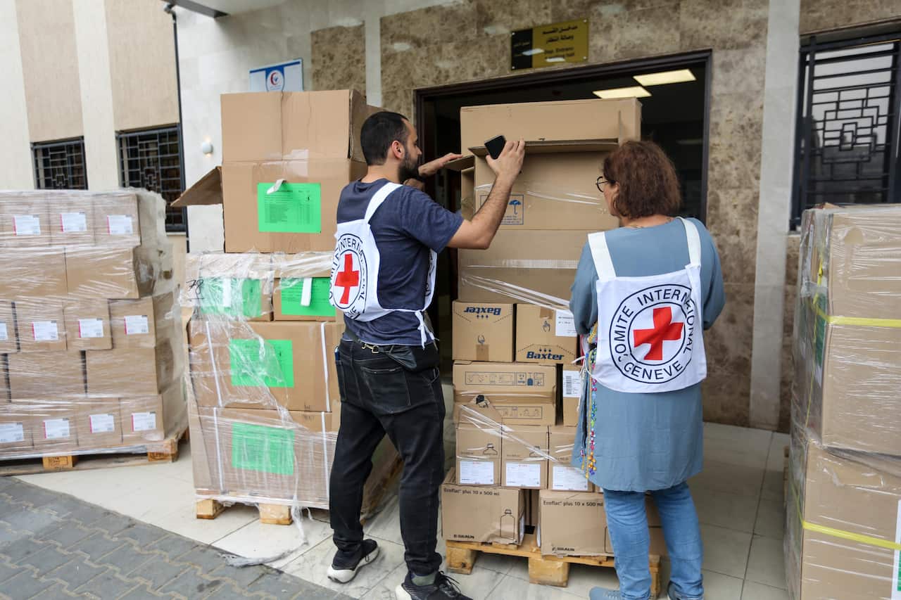 A man and a woman wearing vests with red crosses, inspecting pallets piled high with boxes.