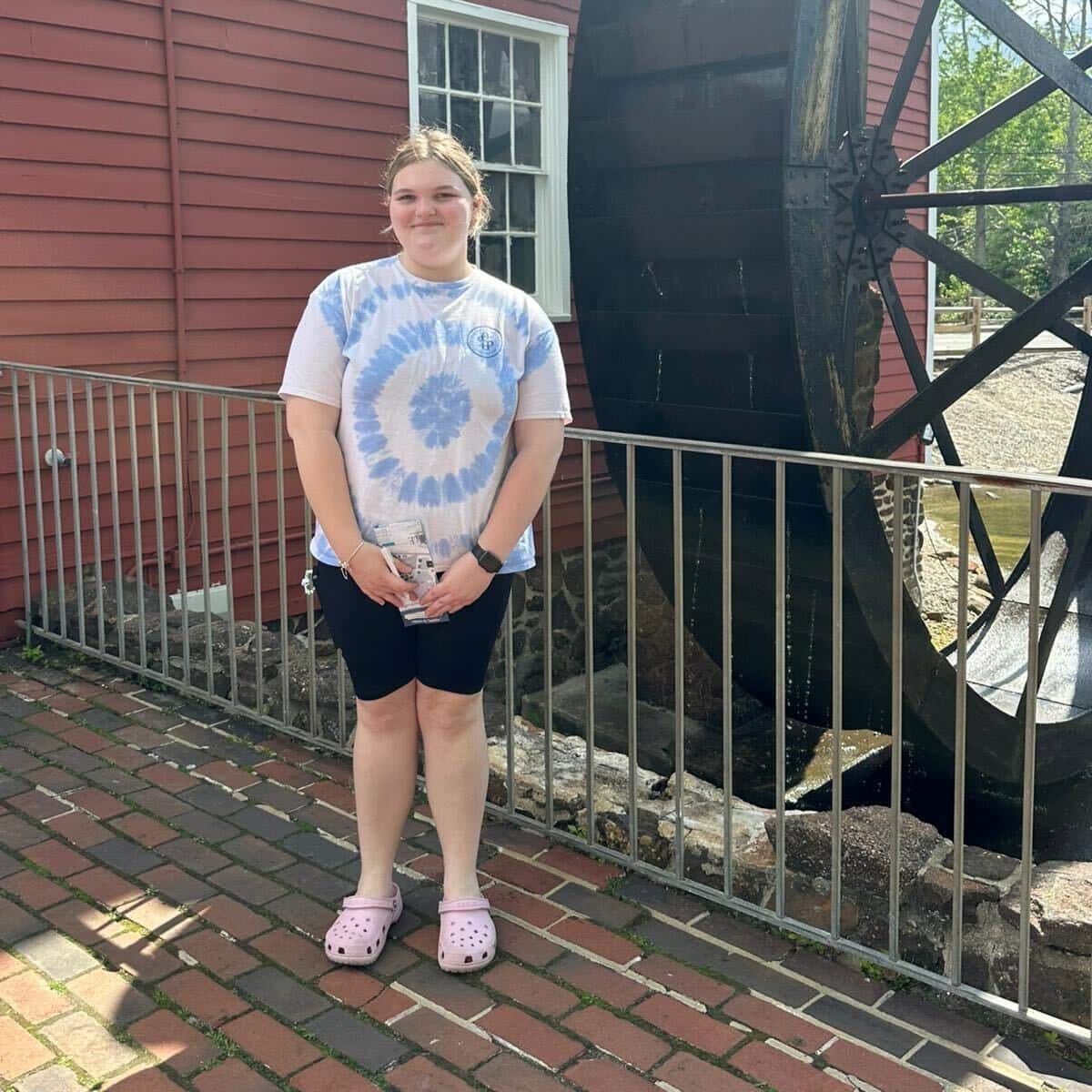 A teenage girl wearing a white-and-blue printed t-shirt and black shorts standing next to a house