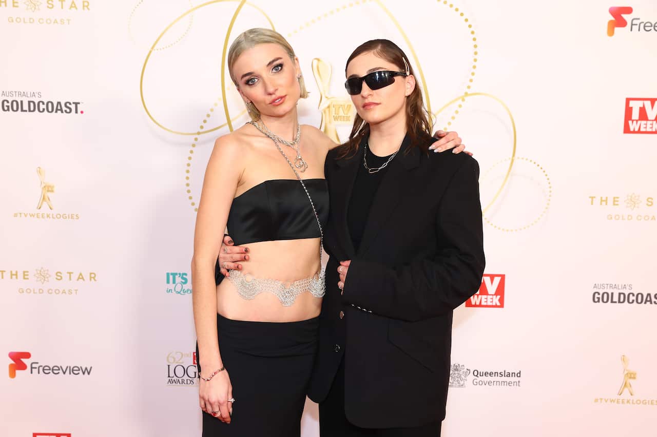 Jenna Owen and Vic Zerbst on a red carpet. Owen has short blonde hair and is wearing a black crop top and long black skirt, with a silver belt around her waist. Zerbst is wearing a black suit and sunglasses.