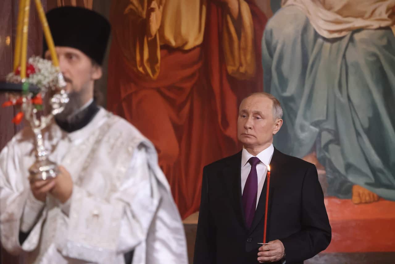 Valdimir Putin, wearing a black suit and red tie, holds a lit red candle as he stands in a church behind a priest wearing white robes.