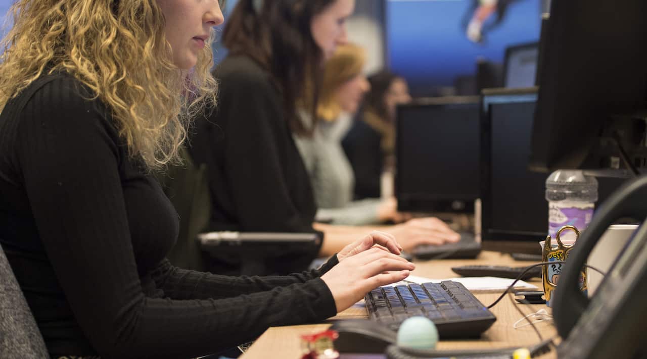 A woman in a black shirt with curly blonde hair sits at a desk in front of a computer in an office. 