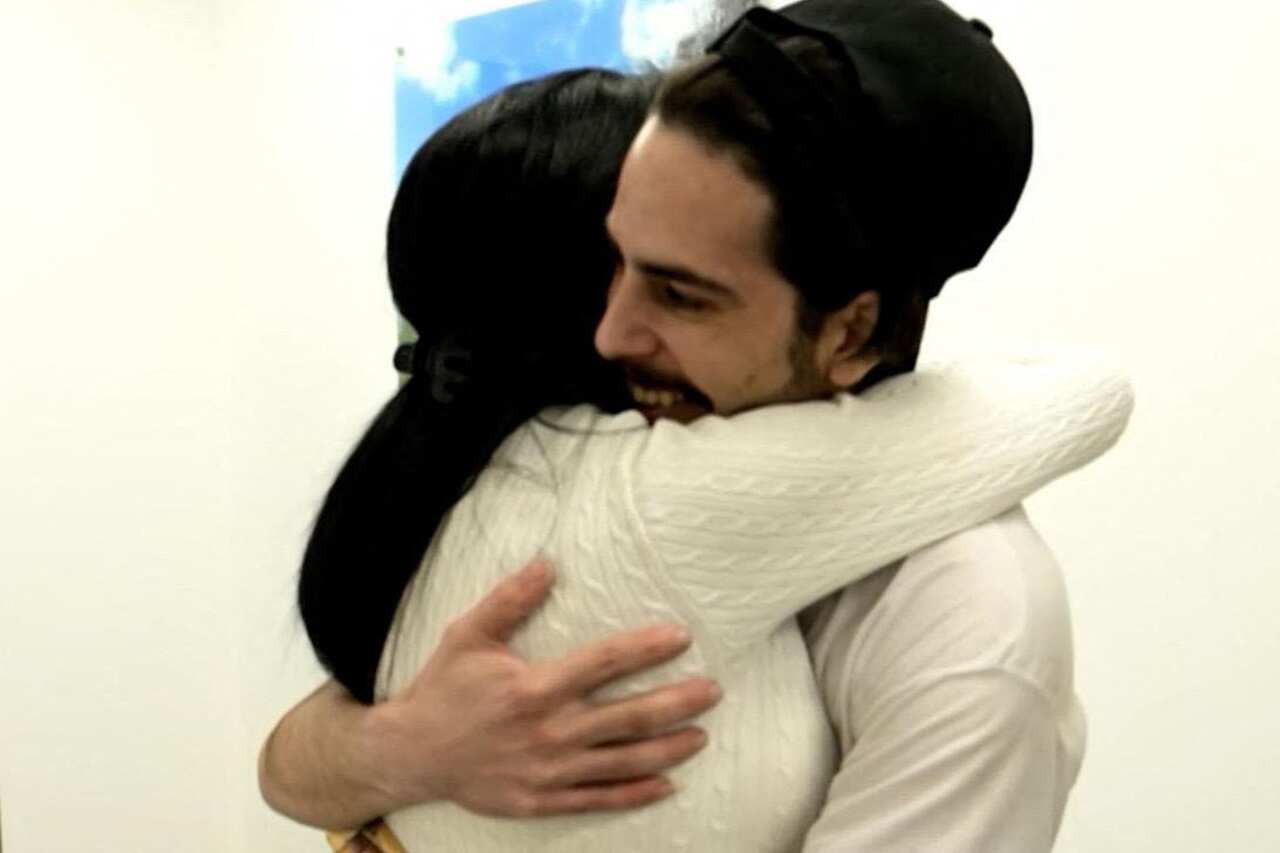 A man in a white shirt and a woman in a white sweater embracing, their arms around one another.