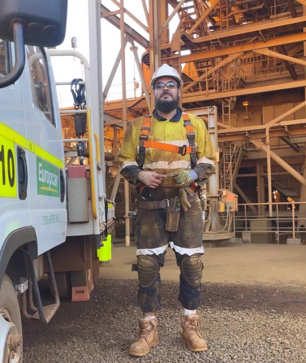 A bearded industrial worker in high-visibility gear, a safety harness, and a hard hat stands outdoors next to a Europcar truck at a mining site.