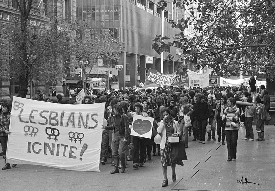 A black and white photo of people walking through Sydney's CBD in the 1970s. At the front people hold a large banner reading Lesbians Ignite