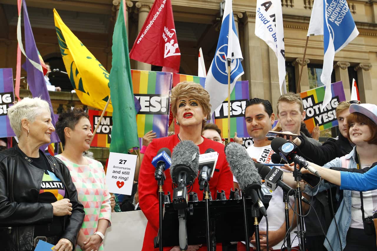 A drag queen addresses the media, surrounded by marriage equality flags and smiling journalists.