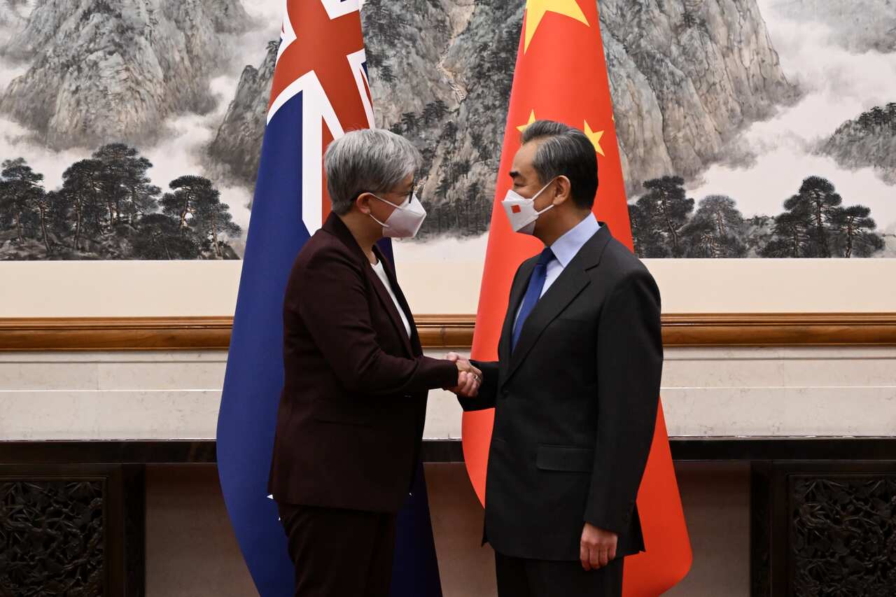 A woman in a dark suit and wearing a face mask (left) shakes hands with a man in a suit and wearing a face mask. There is an Australian flag behind the woman and a Chinese flag behind the man.